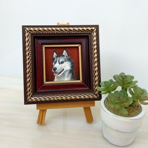 May include: A framed painting of a husky dog with blue eyes. The painting is on a small wooden easel and is in front of a white wall.