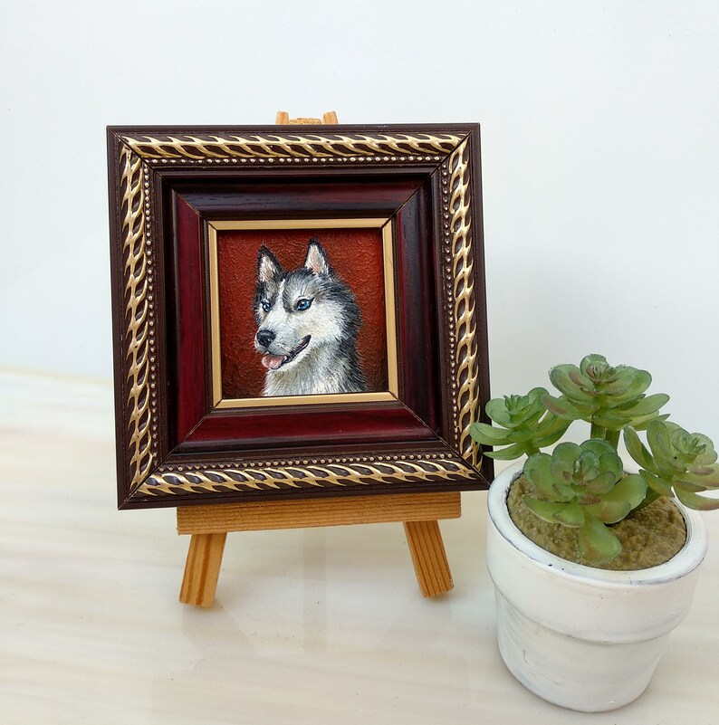 May include: A framed painting of a husky dog with blue eyes. The painting is on a small wooden easel and is in front of a white wall.