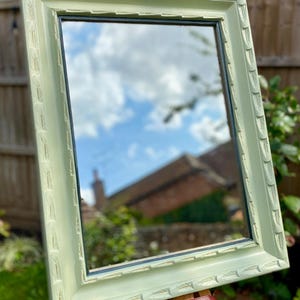May include: A rectangular mirror in a light green, decorative frame. The frame features a repeating pattern. The mirror reflects a cloudy sky and a building. The mirror is propped up on a wooden easel.