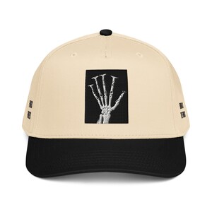 Può includere: Un cappellino da baseball beige con visiera nera e un bottone nero sulla parte superiore. La parte anteriore presenta una toppa rettangolare con una grafica di una mano scheletrica bianca su sfondo nero. Il testo FINGERS FINGERS è stampato sui lati.