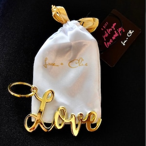May include: Gold-colored metal keychain with the word "Love" in a cursive font. The keychain is packaged in a white satin bag with a gold ribbon and a small brown tag that reads "a gift just for you love and joy's from - Chloe".