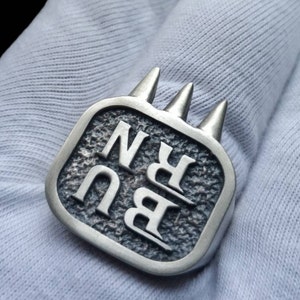 May include: A silver-toned metal ring featuring a square face with a textured, dark grey background. The letters B, U, R, and N are embossed in a bold, serif font. Three pointed, cone-shaped spikes extend from the top edge of the ring face.