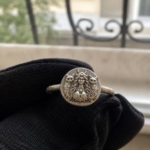 Op de afbeelding: Een zilverkleurige ring met een ronde kop en een reliëfontwerp. Het midden toont een figuur met lang golvend haar en een halve maan als hoofdtooi, geflankeerd door twee katten aan weerszijden. De ringband heeft een getextureerde, verweerde afwerking.