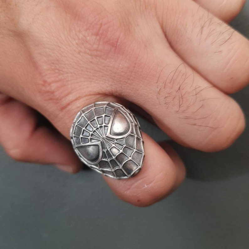 May include: A silver ring with a detailed design of a spider-man mask.