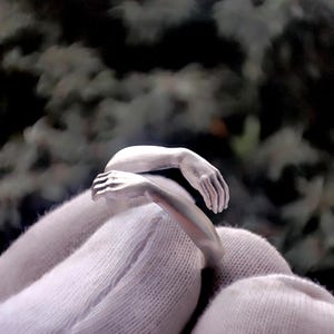 May include: A silver ring shaped like two arms embracing. The ring features detailed hands and arms, creating a unique and artistic design. The ring is shown against a white background.