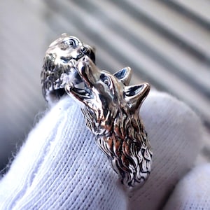 May include: A silver ring with two wolf heads. One wolf is howling, the other is looking at it. The ring is made of a shiny, metallic material. The ring is a piece of jewellery.