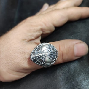 May include: A silver ring with a detailed design of a spider web and a spider's face.
