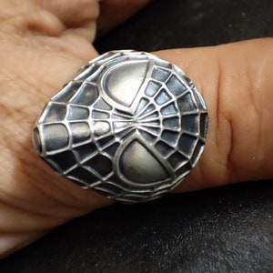 May include: A silver ring with a detailed design of a spider web and eyes. The ring is worn on a finger.