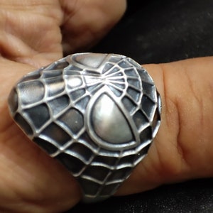 May include: A silver ring with a detailed design of a spider web. The web is raised and textured, and the center of the web features a smooth, rounded shape.