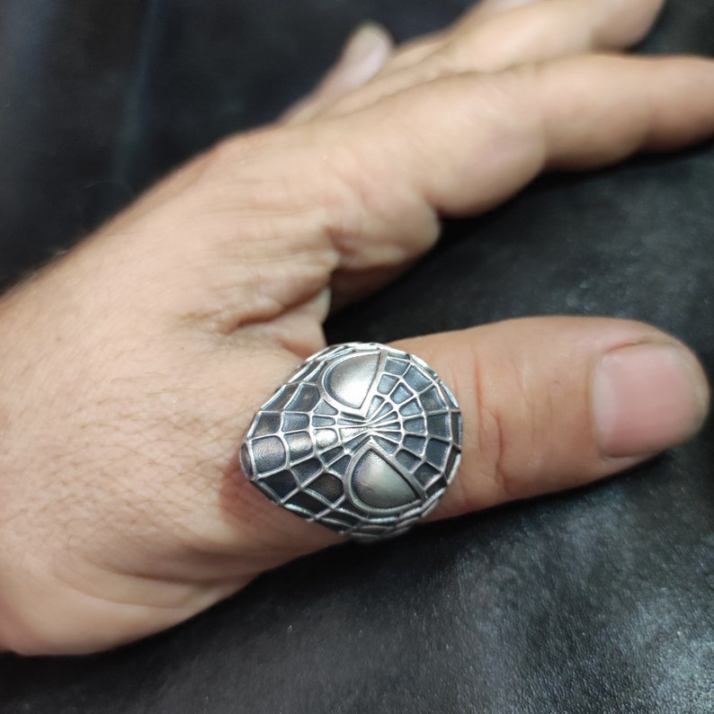 May include: A silver ring with a detailed design of a spider web and a spider's face.