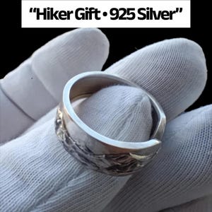 Può includere: Un anello d'argento con un design a catena montuosa. L'anello è in argento 925 ed è etichettato come "Hiker Gift". L'anello è tenuto da un guanto bianco.