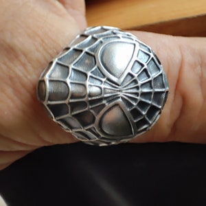 May include: A silver ring with a detailed design of a spider web and a spider's face.