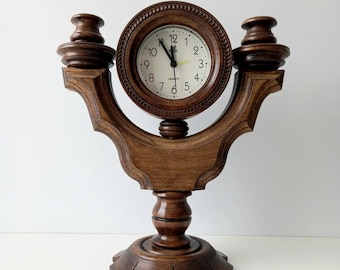 Puede incluir: Un reloj de cuarzo de madera marrón oscuro con esfera redonda y números romanos. El reloj está enmarcado en un marco de madera ornamental y decorativo con base de pedestal y dos remates decorativos. Las manecillas del reloj indican aproximadamente las 11:00.
