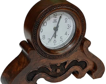 Può includere: Un orologio da mensola in legno marrone scuro con un quadrante bianco. L'orologio ha un quadrante rotondo con numeri e lancette nere. L'orologio è supportato da una base decorativa con dettagli intagliati. Il quadrante recita "PEARL QUARTZ".