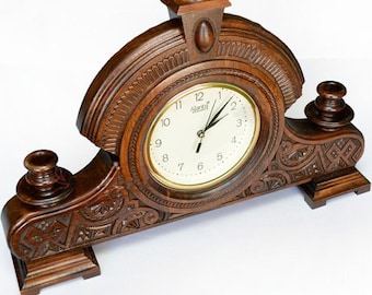 May include: A brown wooden mantel clock with an off-white clock face and gold-colored trim. The clock has ornate carvings and decorative finials. The clock face displays the brand name "Ajanta".
