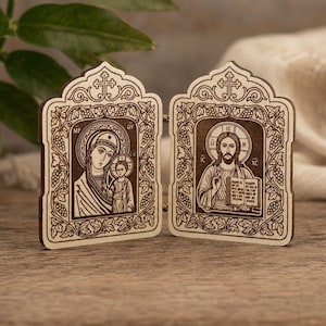 Puede incluir: Un díptico de madera con bisagras que presenta iconografía religiosa grabada con láser. El panel izquierdo muestra una figura con un niño, mientras que el derecho muestra una figura barbuda con un libro abierto, enmarcados por motivos de vides.