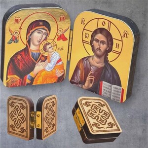 Może przedstawiać: Drewniane pudełka na ikony na zawiasach z wizerunkami religijnymi. Jedno pudełko przedstawia Maryję i Jezusa, drugie Jezusa. Pudełka są złote, czerwone i brązowe z mosiężnymi zawiasami. Pokazano r&oacute;wnież mniejsze pudełko z rzeźbionym kwiatowym wzorem.