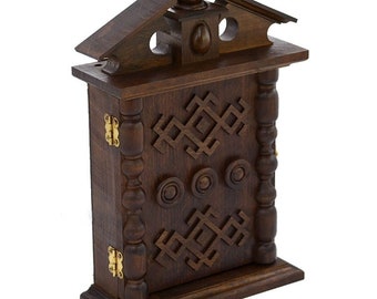 Op de afbeelding: Een donkerbruine houten sleutelhanger met een decoratief gesneden ontwerp. De sleutelhanger heeft een klein dak en een scharnierende deur.