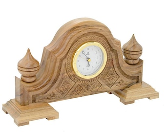 Può includere: Orologio da mensola in legno marrone chiaro. L'orologio ha un quadrante rotondo con finiture dorate e sfondo bianco. L'orologio è decorato con dettagli intagliati e ha due pinnacoli decorativi. L'orologio misura circa 25 cm di larghezza.