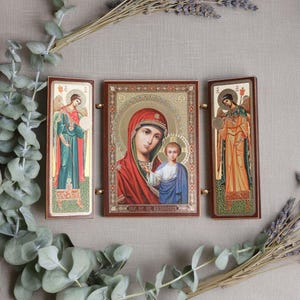 Peut inclure: Un triptyque d'icônes religieuses représentant la Vierge Marie et l'Enfant Jésus, flanqué d'images d'anges. Les icônes ont des accents dorés et sont encadrées de bois foncé. L'icône centrale représente Marie dans un voile rouge.