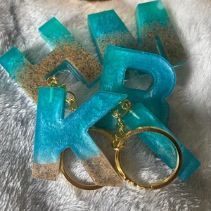 May include: Resin letter keyrings in shades of blue and turquoise, with a sand-like texture at the base. Each letter has a gold-coloured keyring attached. The keyrings are arranged on a white, textured surface. The letters spell out a name.