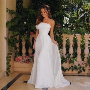 Op de afbeelding: Een vloerlange witte strapless japon met een aansluitend lijfje met gedrapeerde stofdetails en een zwierige, gelaagde rok. De jurk is gemaakt van een lichtgewicht, semi-transparant materiaal dat een zacht en elegant silhouet creëert.
