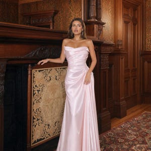 Op de afbeelding: Een vloerlange, strapless lichtroze satijnen jurk met een gedrapeerd lijfje in cowl-stijl en een geplooide taille die overgaat in een gladde, vloerlange rok. De stof heeft een subtiele glans, wat zorgt voor een zachte, reflecterende uitstraling.