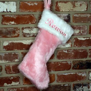 May include: Pink faux fur Christmas stocking with a white faux fur cuff and the name "Rowan" embroidered in pink.