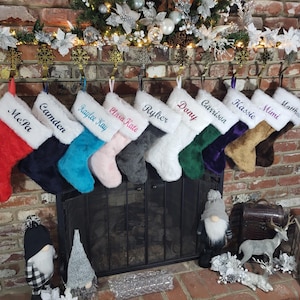 May include: A collection of personalized Christmas stockings in various colors, including red, navy, teal, pink, gray, and white, each with a name embroidered on the white cuff. The stockings are hanging on a brick fireplace, decorated with festive ornaments.