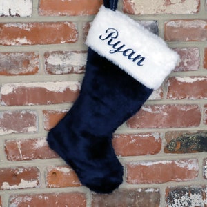 May include: A navy blue Christmas stocking with a white faux fur cuff. The stocking is personalized with the name "Ryan" embroidered in white.