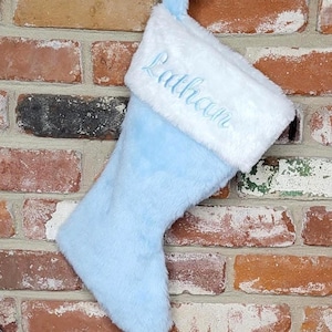 May include: A light blue Christmas stocking with a white cuff. The name "Lathan" is embroidered in light blue script on the cuff. The stocking is hanging on a brick wall.