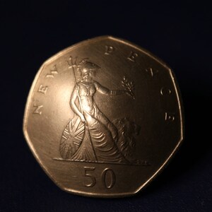Könnte beinhalten: Eine silberne 50-Pence-Münze mit einer Darstellung von Britannia, einer weiblichen Figur, die einen Dreizack und einen Zweig mit Olivenblättern hält, die auf einem Schild mit der Union Jack steht. Die Münze ist mit "NEW PENCE" und "50" beschriftet.