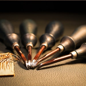 May include: A set of six leather working tools with black handles and red metal blades. The tools are arranged in a fan shape on a brown leather surface.