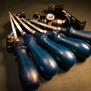 May include: Six leather working tools with blue handles and silver blades. The tools are arranged in a fan shape on a brown leather surface.