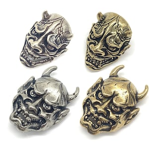 Könnte beinhalten: Vier verschiedene farbige japanische Oni-Masken aus Metall. Die Masken haben alle das gleiche Design, sind aber in Gold, Silber, Messing und dunklem Messing ausgeführt.