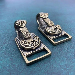 May include: Two ornate brass belt buckles with intricate designs. The buckles have a hook on the top and a rectangular shape. The buckles are a dark brass color with gold accents.