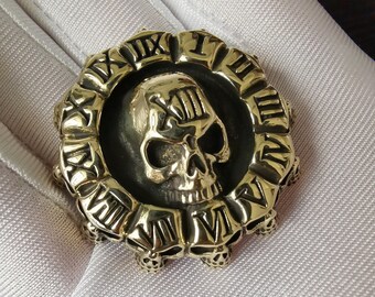 May include: A gold-toned ring with a skull design in the center. The skull is surrounded by a circle of Roman numerals. The ring is set on a white background.