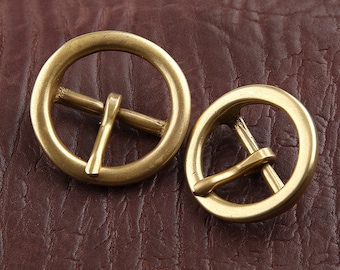 May include: Two brass-coloured, round belt buckles with a central bar and a pin. The buckles are of different sizes, with a larger one on the left and a smaller one on the right. They are set against a textured, brown leather background.
