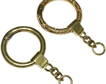 Könnte beinhalten: Zwei goldene Schlüsselanhänger mit verschiedenen Ringformen. Ein Ring ist schlicht, der andere hat ein verziertes Design. Beide Schlüsselanhänger haben eine Kette mit einem kleinen Ring am Ende.