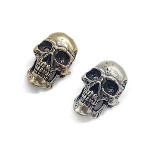 Pode incluir: Dois botões de metal em forma de caveira, um dourado e outro prateado. As caveiras são detalhadas com dentes e órbitas.
