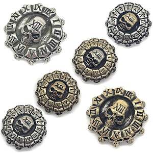 Puede incluir: Cinco abalorios redondos de metal con forma de calavera y esfera de reloj. Los abalorios son de color plateado y dorado. Las esferas de reloj tienen números romanos y una calavera en el centro. Los abalorios están rodeados por un anillo de pequeñas calaveras.