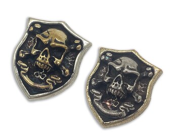 May include: Two silver and gold skull and crossbones shield-shaped buttons. The skulls are detailed with teeth and eye sockets. The crossbones are stylized and have a wing-like design.