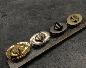 May include: Four oval-shaped metal clasps in different finishes: gold, silver, black, and brass. The clasps are arranged in a row on a textured brown surface.