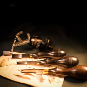 May include: Four wood carving tools with dark wood handles and brass accents. The tools are lying on a piece of light brown leather.