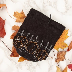 May include: A black drawstring bag with an embroidered design of pumpkins and a fence. The bag is surrounded by autumn leaves in shades of red, orange, and yellow.