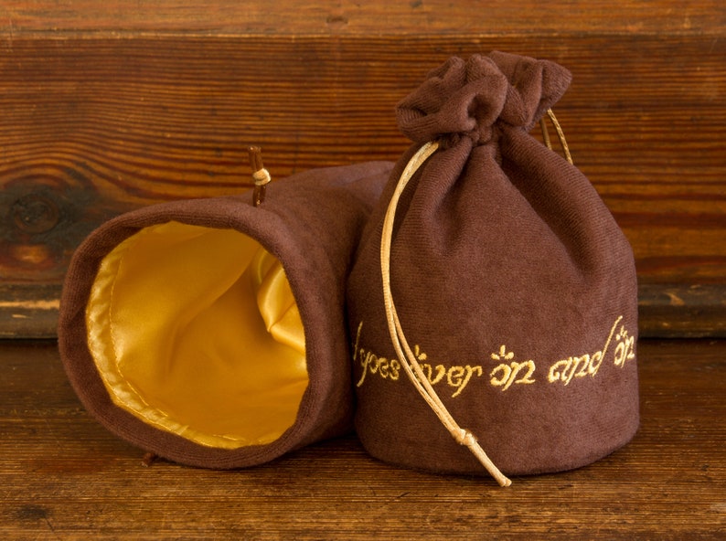May include: Two brown velvet pouches with gold drawstring closures. One pouch has a gold satin lining and the other has the inscription "One Ring to rule them all, One Ring to find them, One Ring to bring them all and in the darkness bind them" embroidered in gold thread.