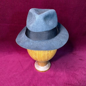 Może przedstawiać: Szary kapelusz typu fedora z czarną wstążką. Kapelusz jest prezentowany na drewnianym stojaku. Kapelusz ma klasyczny, ponadczasowy design. Rondo kapelusza jest lekko zakrzywione, a korona wklęsła.