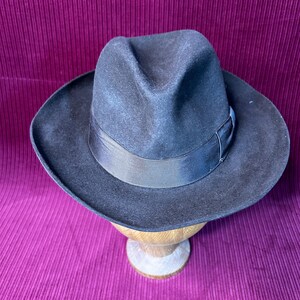 May include: A black fedora hat with a wide brim and a black ribbon band. The hat is displayed on a wooden hat stand against a deep purple background. The hat has a classic, timeless design.
