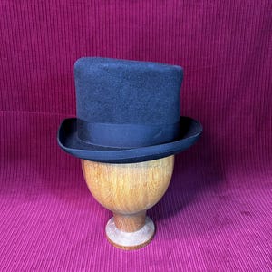 Peut inclure: Un chapeau haut de forme bleu marine foncé avec un ruban noir, présenté sur un support en bois. Le chapeau a une couronne haute classique et un large bord. Le fond est un tissu rose foncé texturé.