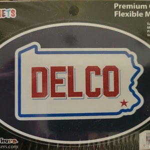 Peut inclure: Un aimant ovale bleu et blanc avec le mot "DELCO" en lettres rouges et une étoile rouge. L'aimant est en forme de l'état de Pennsylvanie. L'aimant est sur un fond noir avec les mots "Premium Quality Flexible Magnet" et "To Remove Magnet Push Out From Inner Cut Line" en texte blanc.